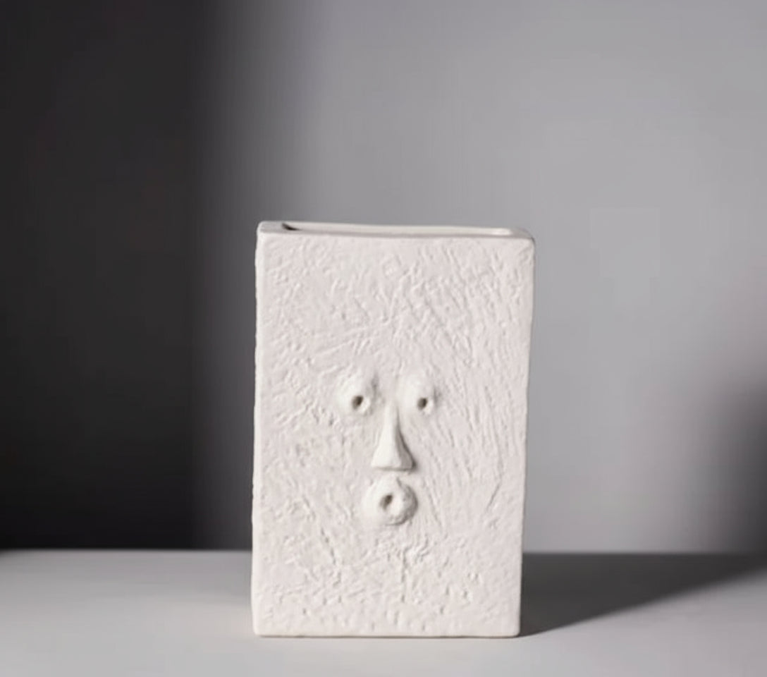 A white ceramic vase with an abstract face design, displayed on a table with a grey background.